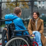 Woman and man in wheelchair conversing outdoors, smiling, urban background